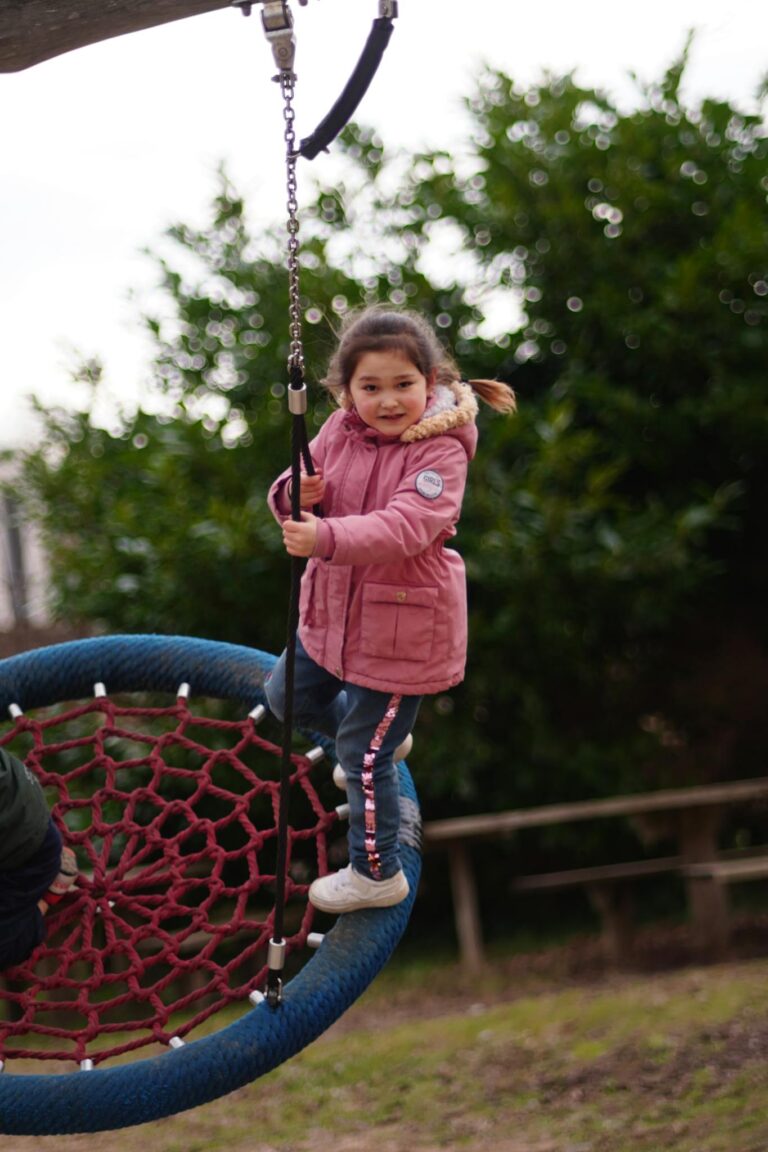 A joyful child in a pink coat swings at a lush green playground on a cloudy day.
