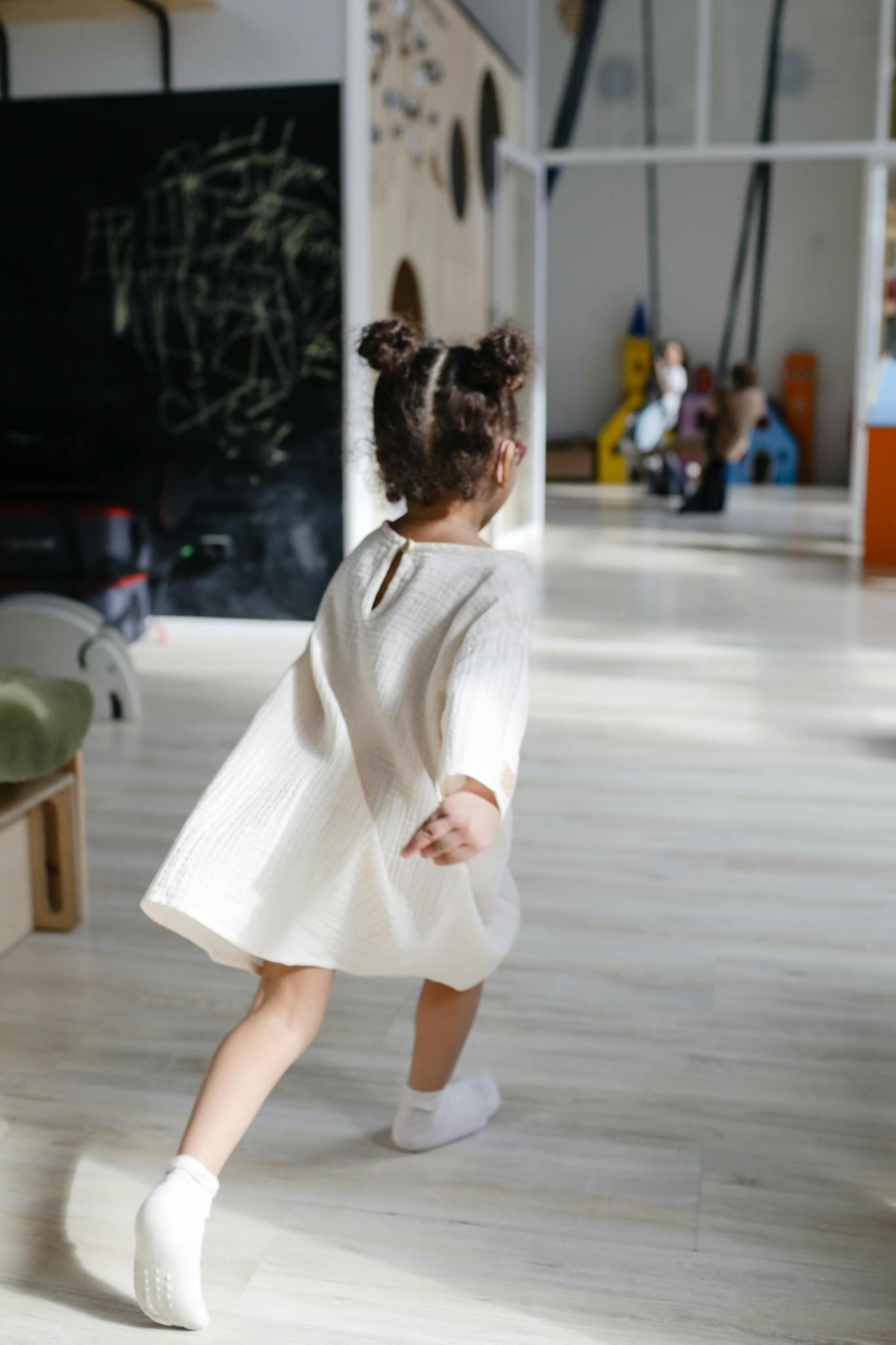 A young girl with hair buns runs joyfully in a sunlit playroom wearing a white dress.