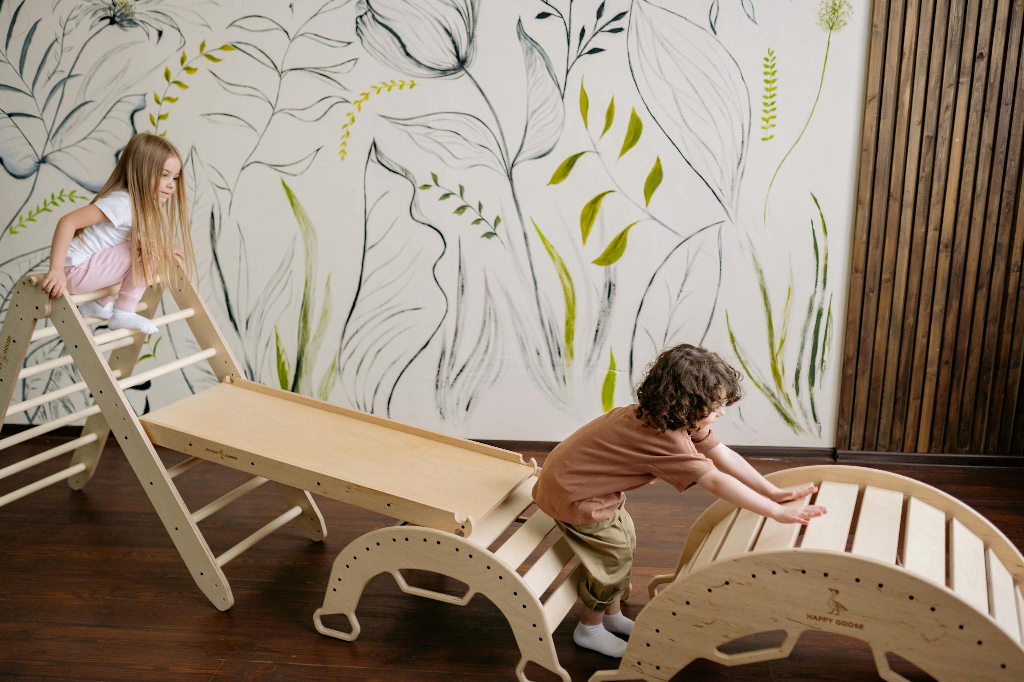 Children enjoying playtime on a wooden slide in an indoor playroom with artistic wall designs.